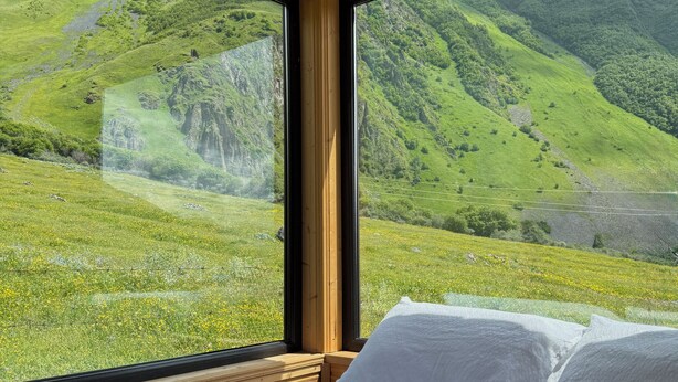 A bedroom with a large window offering a view of a green valley and mountains. - Kazbegi Inn Cottages  (Kazbegi)