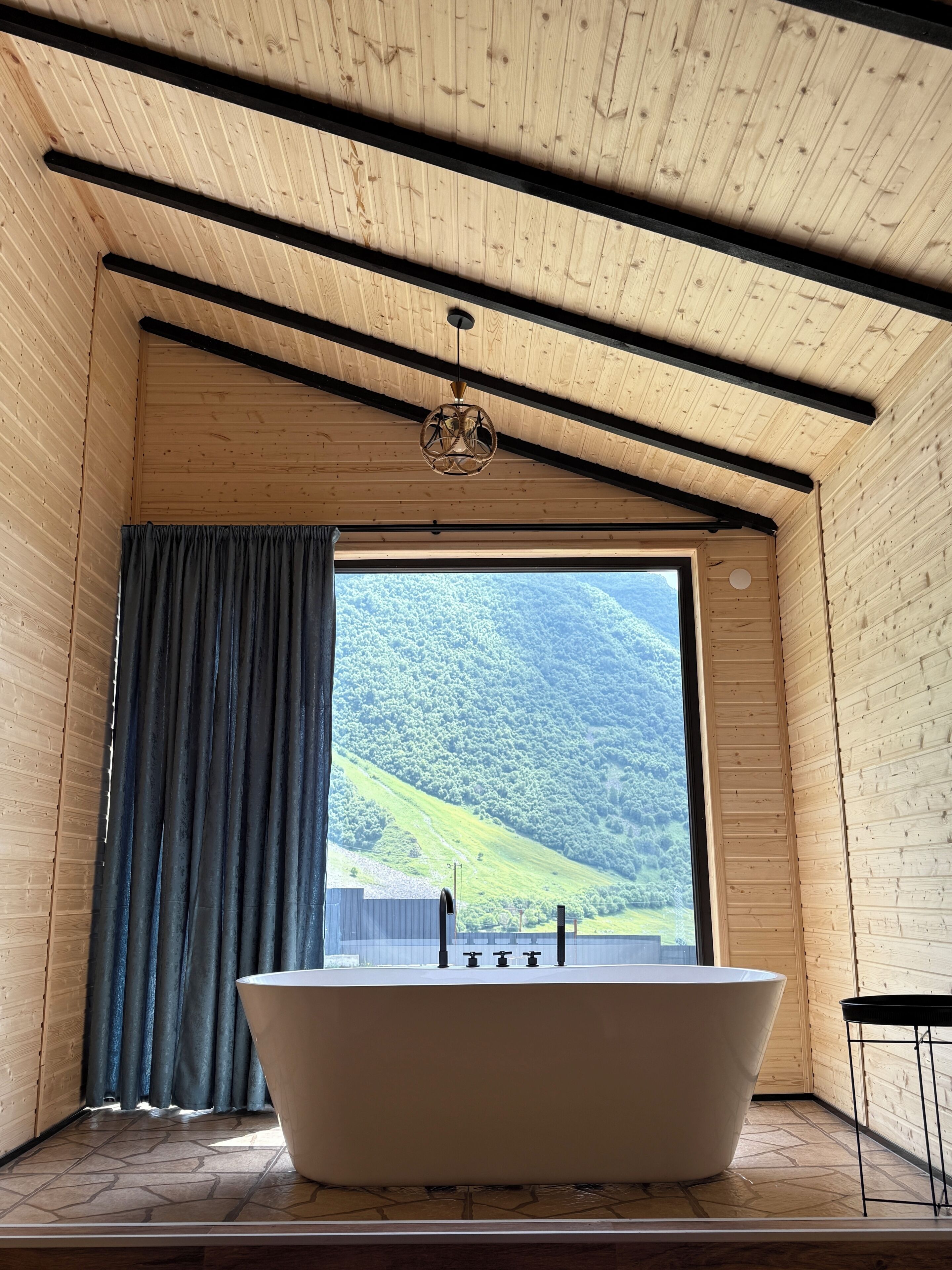A room with a large bathtub, wooden walls, a view of a green hillside, and a curtain.