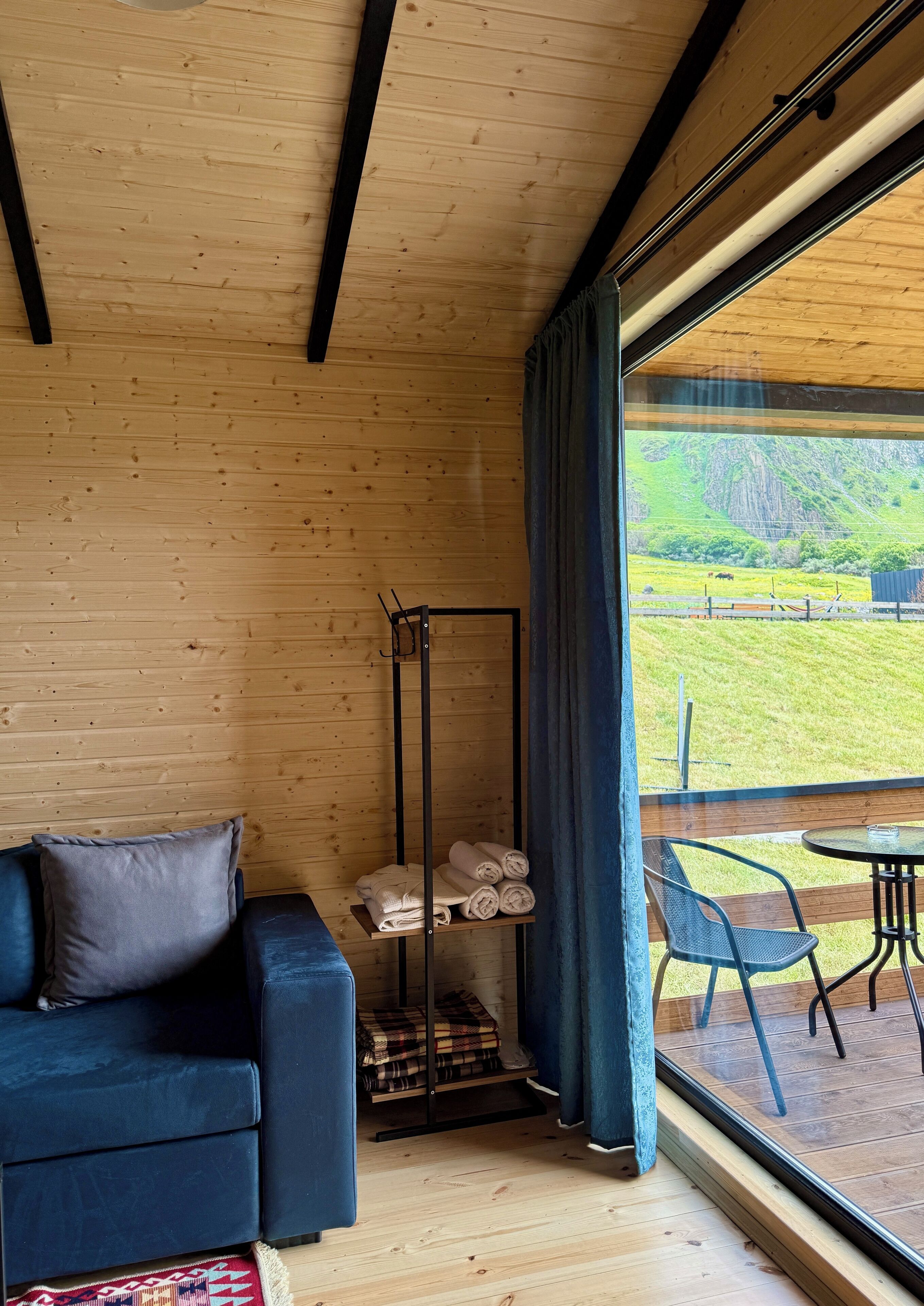 Een kamer met een houten plafond, een blauwe bank, een metalen rek met handdoeken en uitzicht op een groen landschap door een groot raam.