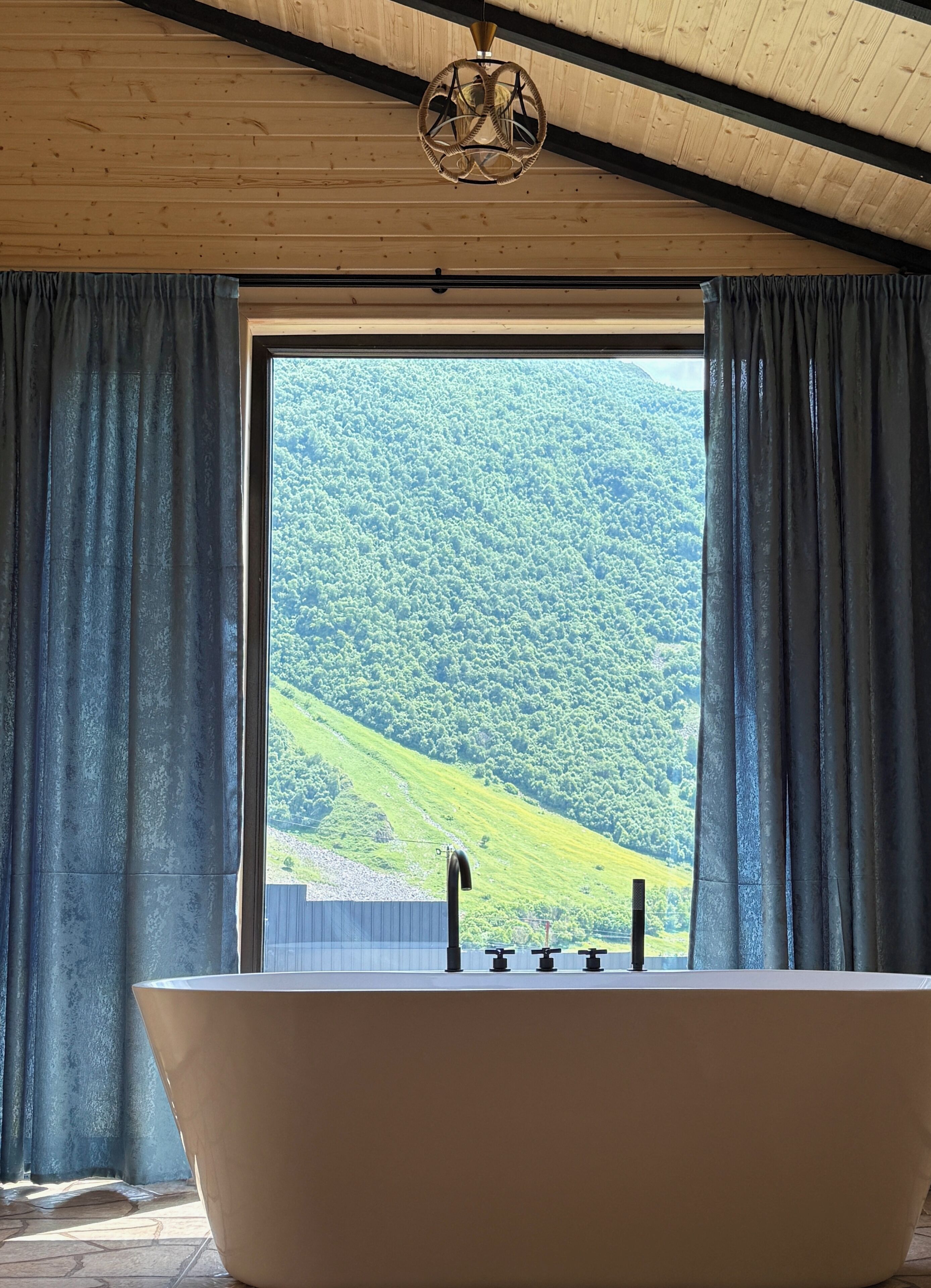 Un baño con una bañera independiente, techo de madera y un amplio ventanal con cortinas.