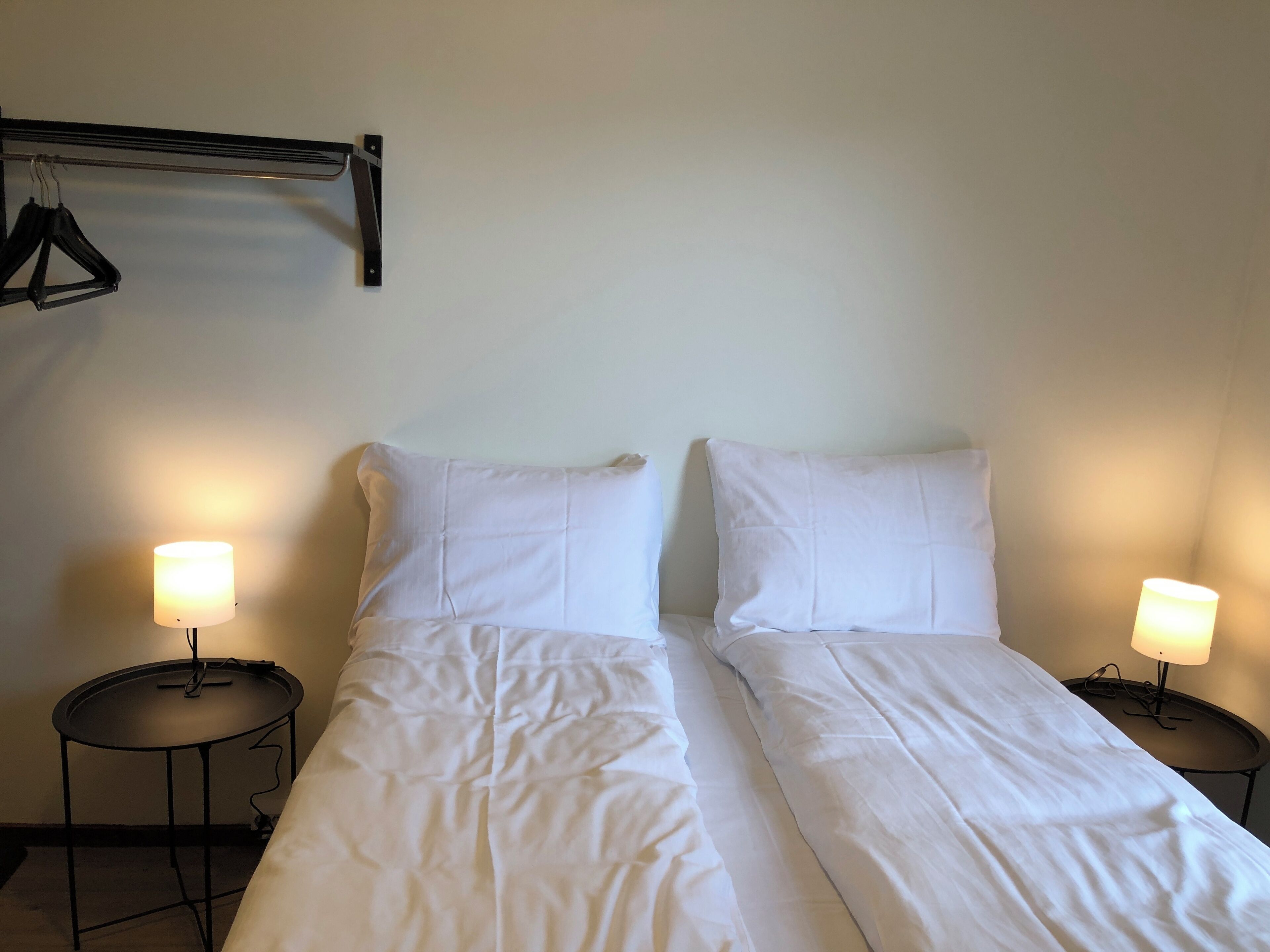 A double bed with white linens, two bedside tables with lamps, and a wall-mounted rack with hangers.