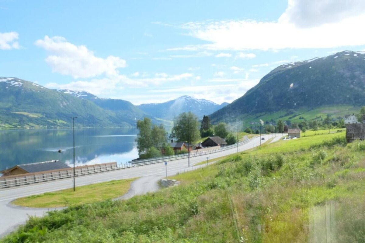 A lakeside road with a bridge, houses, and mountains in the background.