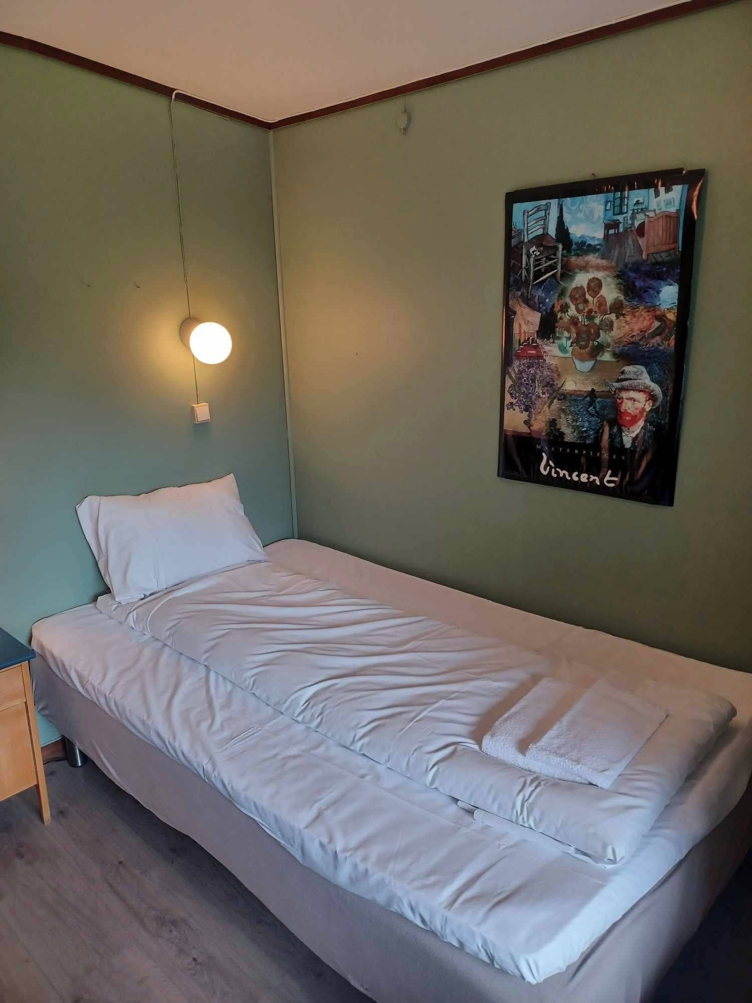 A single-bed room with a green wall, a painting, and a bedside lamp.