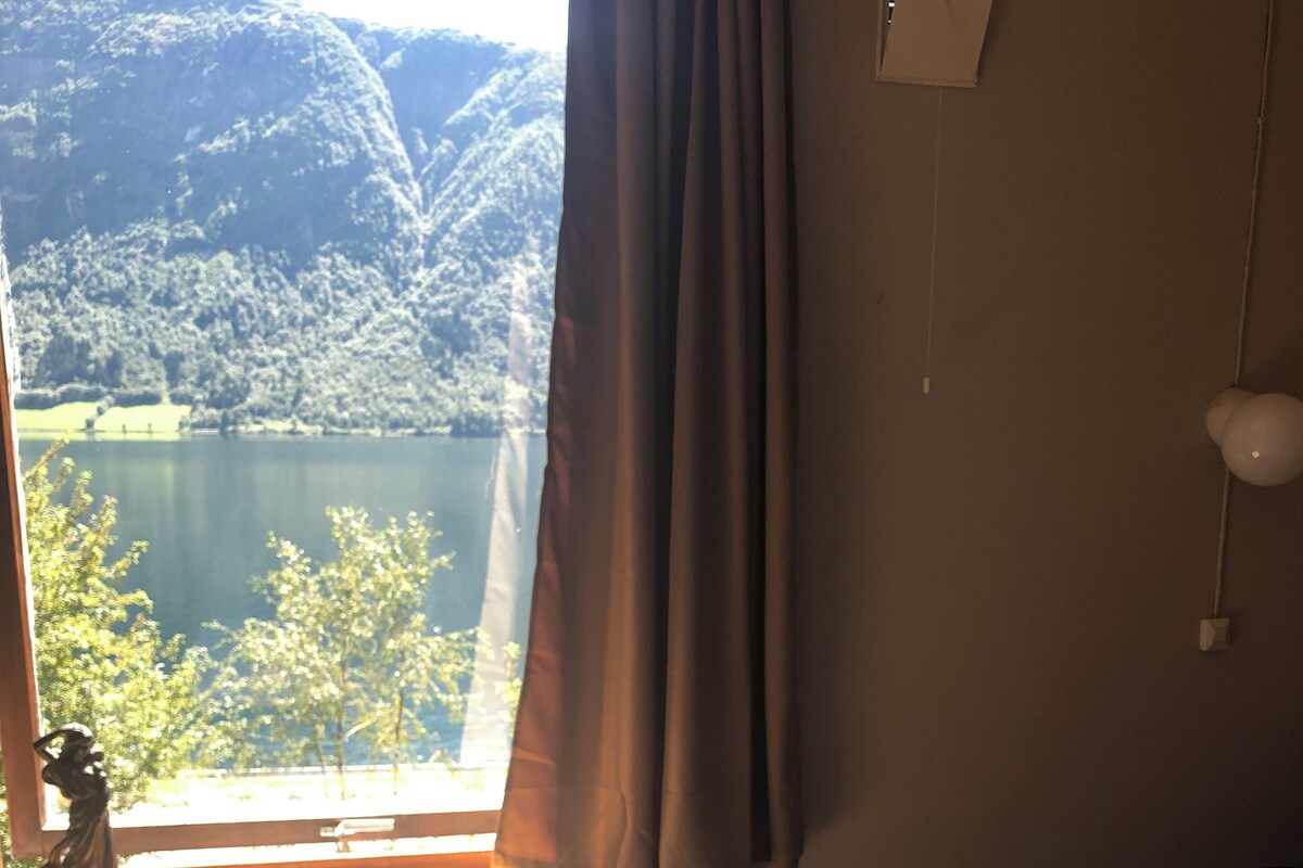A hotel room with a bed, a window with curtains, a radiator, a wall-mounted light fixture, and a view of a lake and mountains.