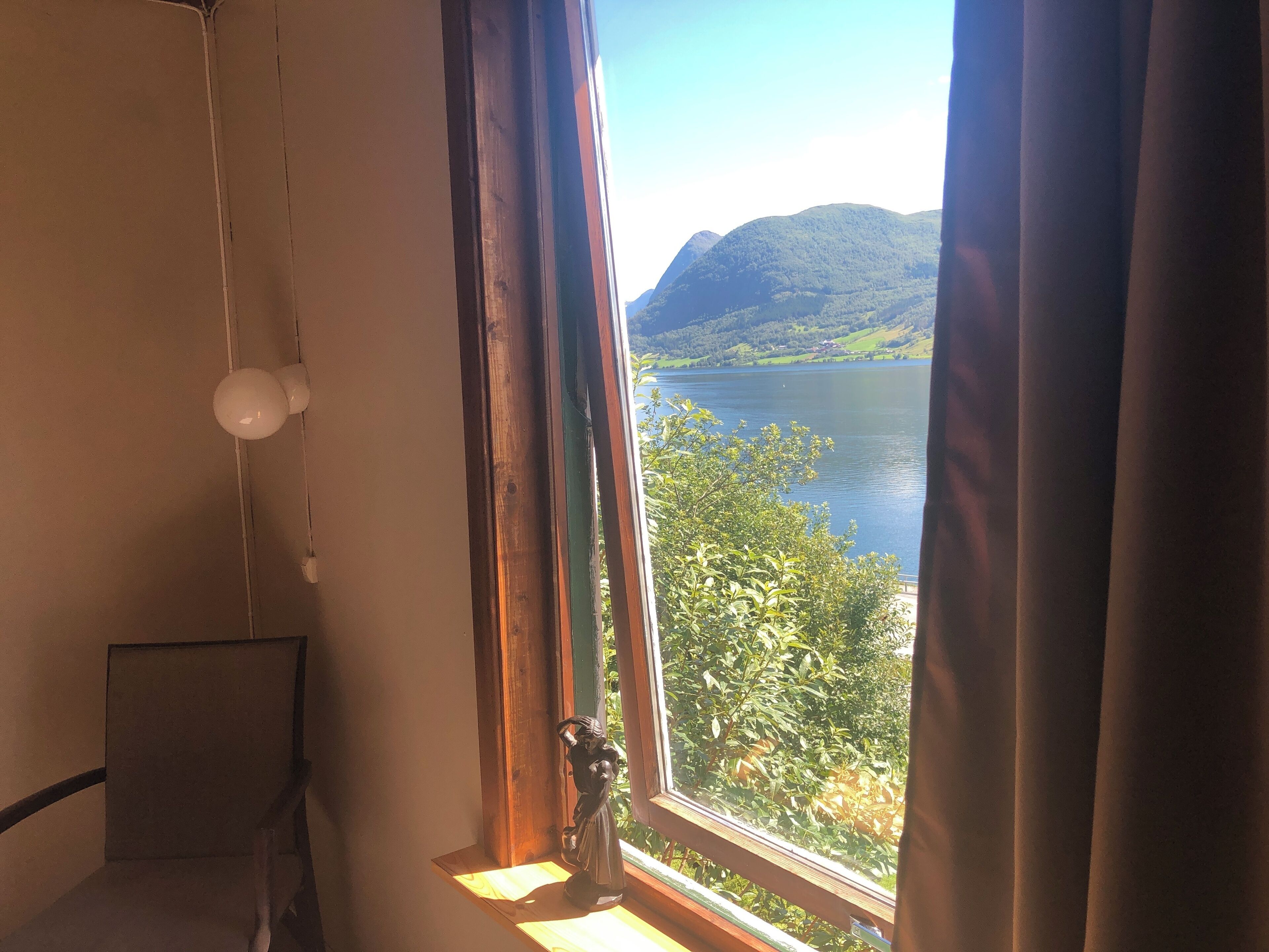 A room with a large window overlooking a lake and hills. There is a chair and a small statue on the windowsill.