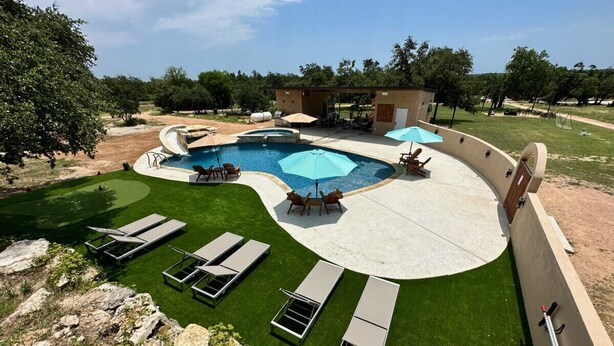 A swimming pool with a curved design, surrounded by a concrete deck and a grassy area with lounge chairs. - The Family Retreat in Dripping Springs (Dripping Springs)