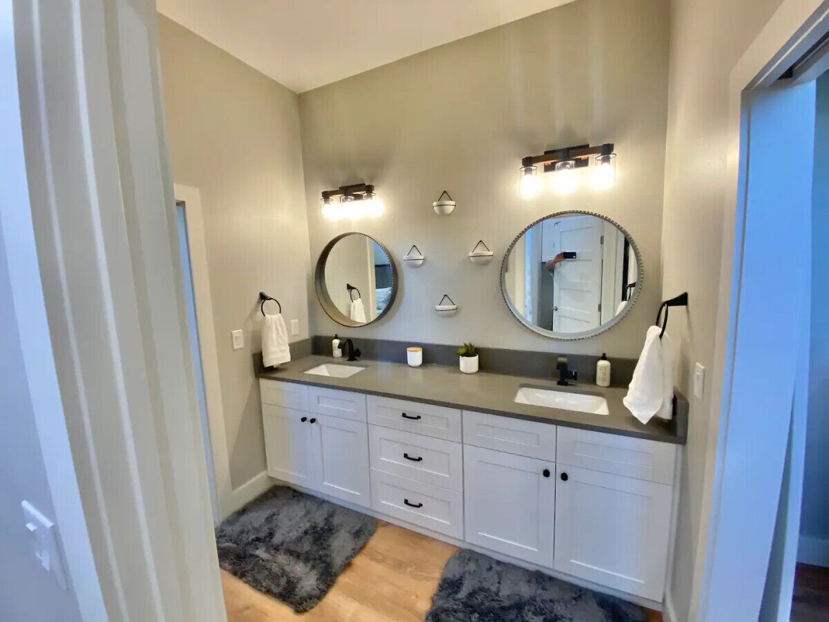 Un bagno con due lavandini, un mobile lavabo e due specchi fissati alla parete. Sopra gli specchi sono presenti dei corpi illuminanti e un asciugamano è appeso sul lato destro.