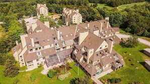 A large, multi-story hotel with a central courtyard, surrounded by lush greenery and a golf course in the background. - BaseLoj - Pool, Firepit, Bike to Mountain! (Killington)