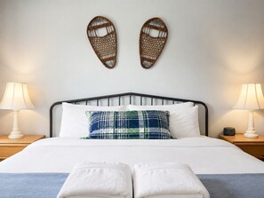 A neatly made bed with white linens, a plaid pillow, and two towels on the bed. There are two bedside lamps on wooden nightstands. - BaseLoj - Fireplace, Hot tub, Pool, Sauna! (Killington)