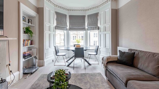 A modern living room with a bay window, a round dining table with chairs, a sofa, and a bookshelf.