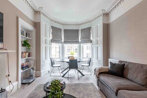 A modern living room with a bay window, a round dining table with chairs, a sofa, and a bookshelf. - Spacious 3 bed home in Marchmont  (Edinburgh)