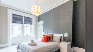 A modern bedroom with a large bed, a bedside table, a lamp, and a view of buildings through a window.