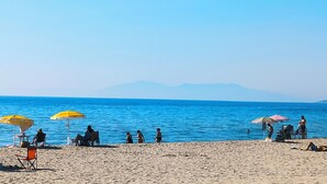 Una playa con gente descansando bajo sombrillas, cielo despejado y montañas al fondo.