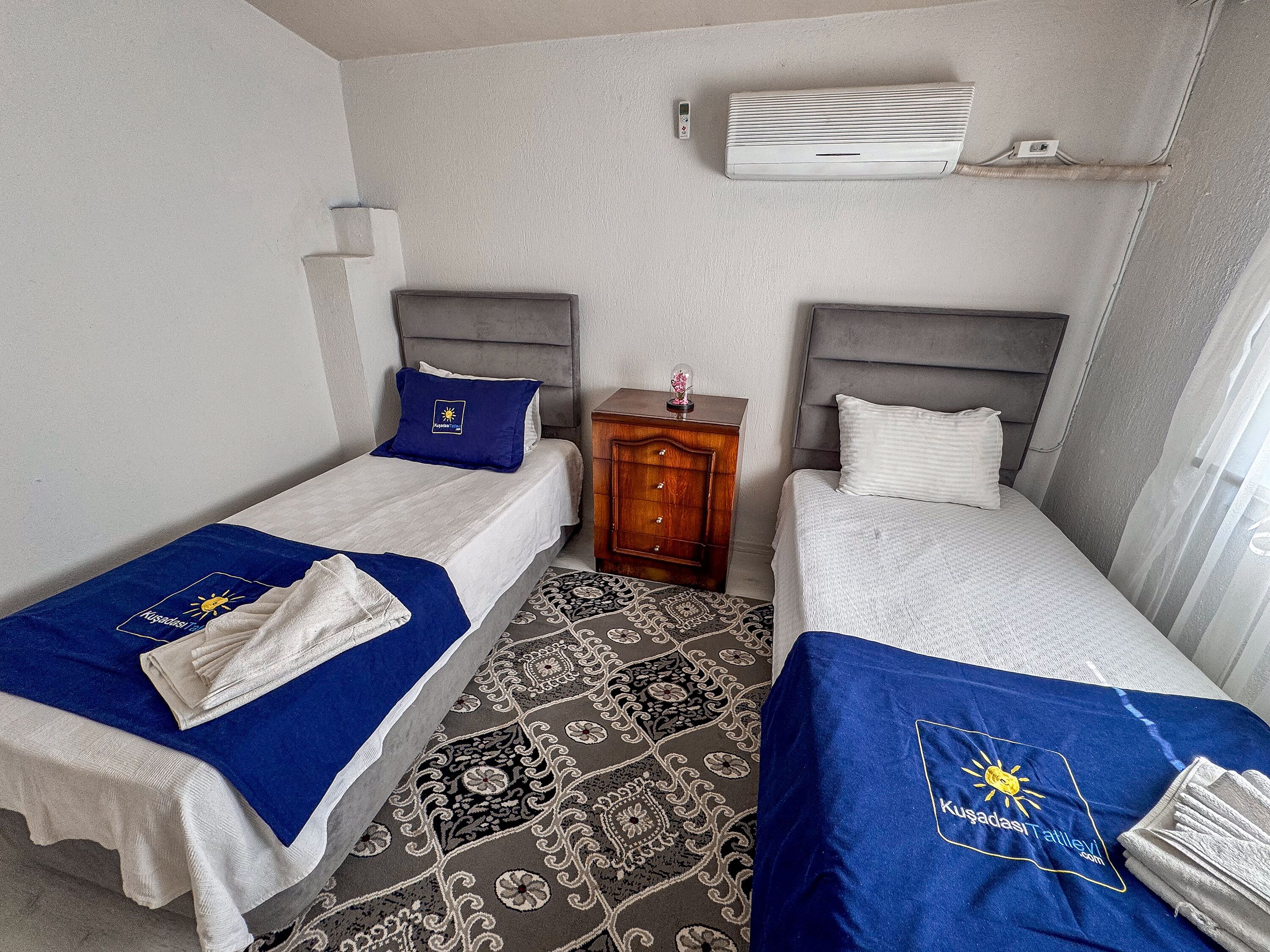 Ein Hotelzimmer mit zwei Betten, jeweils mit einem blauen Handtuch und einem Kopfkissen mit einem Logo. Ein kleiner Nachttisch mit Lampe und einem dekorativen Gegenstand steht daneben. Eine Klimaanlage ist an der Wand oberhalb der Betten angebracht.