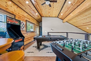 Une pièce aux murs et au plafond en bois, avec une table de billard, une table de baby-foot et une table de baby-foot équipée d’un écran diffusant un match.