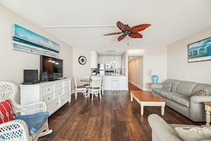 A living room with a TV, white furniture, a dining area, and a kitchen in the background. - Includes Xplorie Activities & Linens. PYRAMID-2BR OF condo w/outdoor pool. (Ocean City)