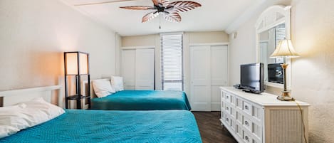 Habitación de hotel con dos camas, ventilador de techo, televisor y cómoda.