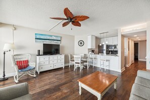 A living room with a television, white furniture, a ceiling fan, and a kitchen area in the background.