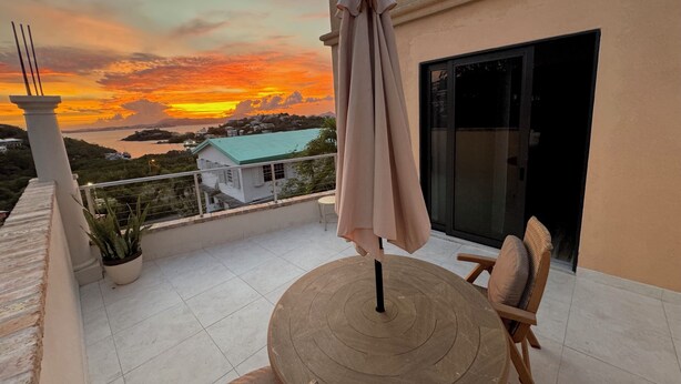 A balcony with a round table and chairs, a potted plant, and a view of a sunset over a body of water. - Sunset Hideaway Suite - Oceanview/central to Cruz Bay  (St John)