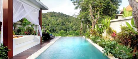 A rectangular swimming pool with a white canopy and steps leading down to a seating area, surrounded by lush greenery and a hilly backdrop.