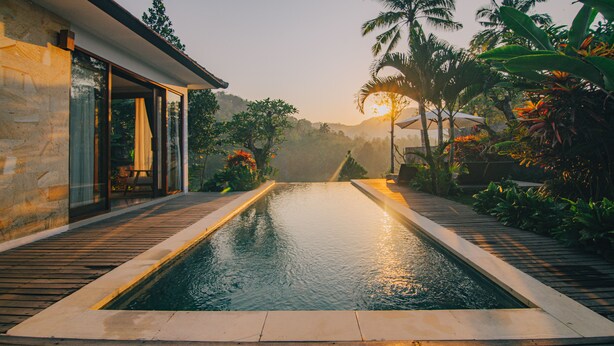 A swimming pool with a wooden deck, surrounded by tropical plants and palm trees, leading to a modern building with large glass windows. - The Sidemen Villas (Sidemen)