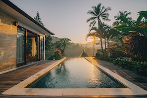 A swimming pool with a wooden deck, surrounded by tropical plants and palm trees, leading to a modern building with large glass windows. - The Sidemen Villas (Sidemen)
