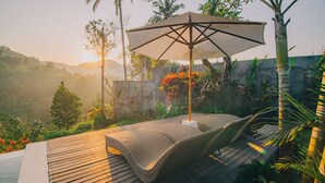A wooden deck with wicker lounge chairs and a large umbrella, surrounded by tropical plants and a view of distant hills. - The Sidemen Villas (Sidemen)