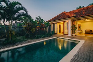 Une piscine avec une terrasse en bois, entourée de plantes tropicales et une maison avec un toit en tuiles.
