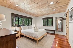 A bedroom with a wooden ceiling, large windows, a bed, a bedside table, and a mirror.