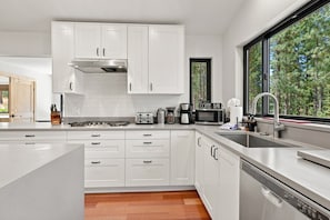 Cozinha moderna com armários brancos, eletrodomésticos de aço inoxidável e vista para a floresta por uma grande janela.