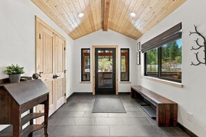 A hallway with a wooden ceiling, a bench, a door leading outside, and a window with a view of greenery.