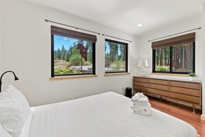 Ein Schlafzimmer mit einem großen Bett, zwei Fenstern mit Jalousien, einer Nachttischlampe, einem Holzk dresser und Blick auf Bäume und ein Gebäude.