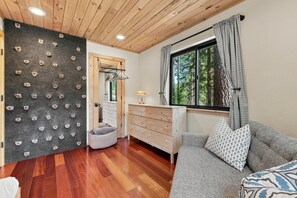 A room with a climbing wall, a window with curtains, a sofa with patterned cushions, and a wooden dresser.