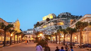 A historic city square with a prominent hilltop building, street lamps, and parked cars.