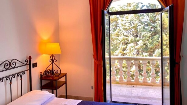 A bedroom with a bed, a bedside lamp, and a view of a balcony with trees.