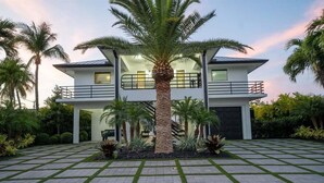 Une maison moderne à deux étages, dotée d’une cour intérieure et entourée de palmiers, ainsi que d’un chemin piétonnier dallé.