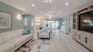 Un salon spacieux avec un canapé blanc, une table basse et un téléviseur posé sur un meuble en bois. On y trouve également un ventilateur de plafond et des spots encastrés.