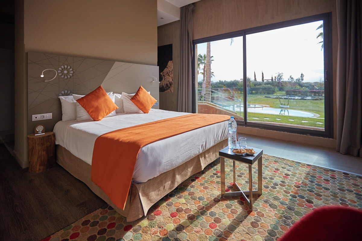 Ein Hotelzimmer mit einem großen Bett, einem kleinen Tisch mit einem Glas Wasser und Blick auf einen angelegten Garten durch ein großes Fenster.