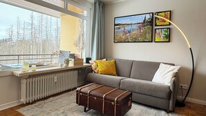 A living room with a grey sofa, a brown trunk, and a window with a view of trees and a building. - Ihr Perfekten Rückzugsort, um die Schönheit des Oberharzes zu Entdecken (Sankt Andreasberg)