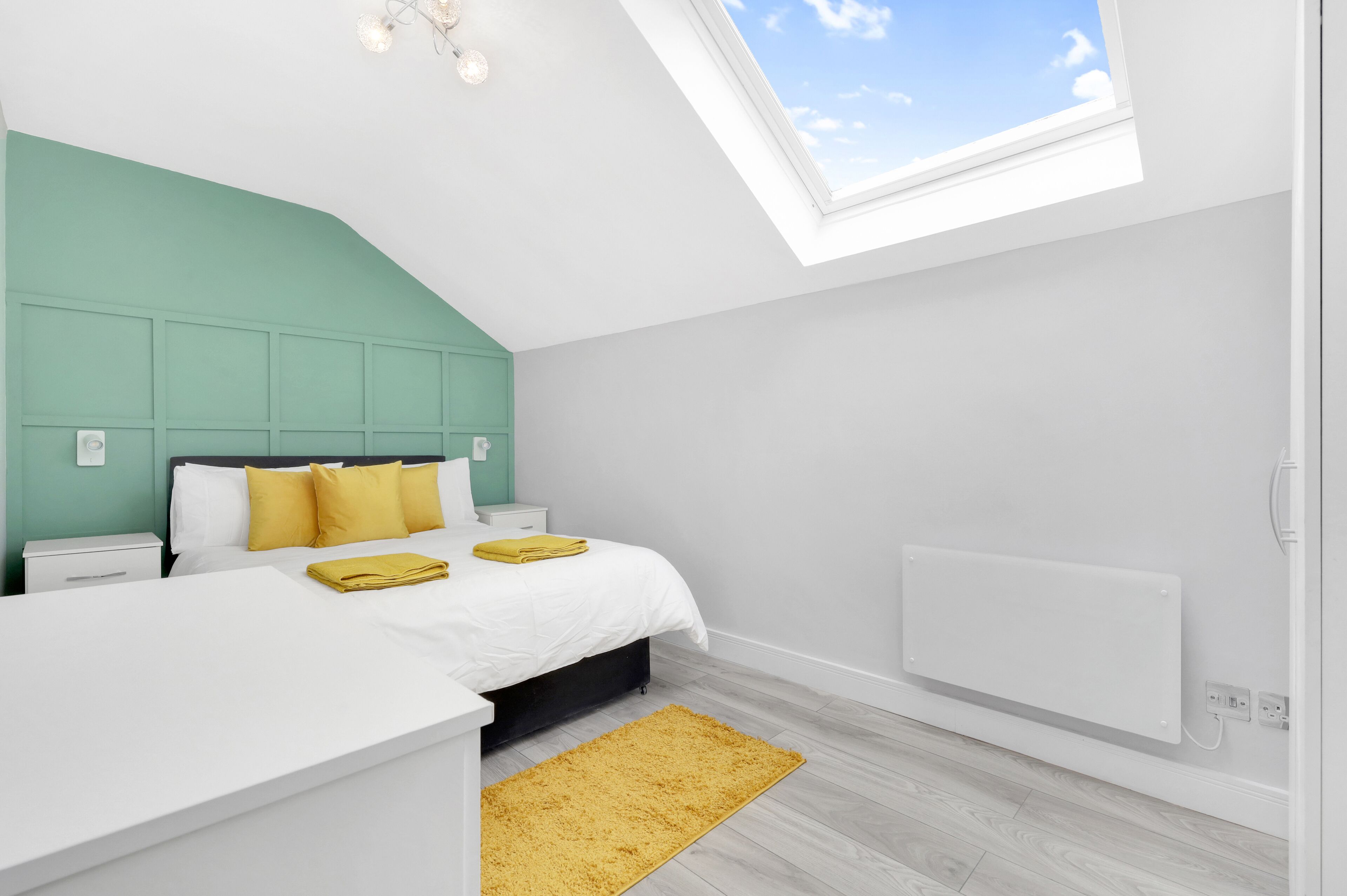 Un dormitorio moderno con tragaluz, una cama con ropa de cama blanca y almohadas amarillas, una mesita de noche blanca y un radiador.