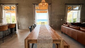 A spacious room with a long wooden table, a leather sofa, and a view of the sea through large windows. - Paradise Cove 3 Bedroom Home (Gregory Town)