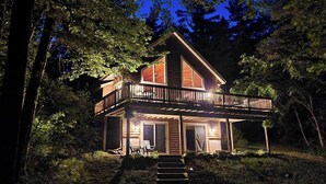 A two-story wooden house with a balcony, surrounded by trees and a wooden pathway leading to the entrance.