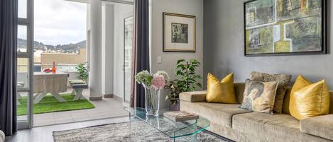 Una sala de estar moderna con una mesa de centro de vidrio, un sofá color beige con cojines dorados, y un cuadro abstracto grande en la pared.