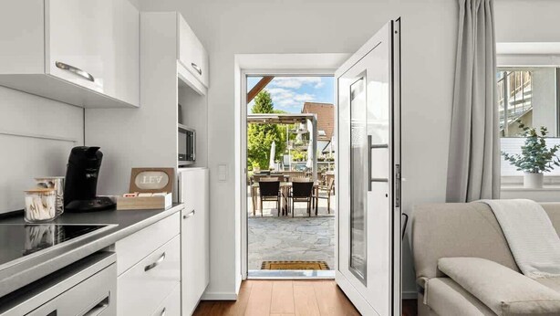 Una cocina moderna con armarios blancos, una encimera negra y una cafetera. Hay una puerta corrediza de vidrio que da a un patio exterior con una mesa y sillas.