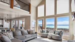 A spacious living room with a large gray sectional sofa, a round gray armchair, and a matching ottoman. The room features large windows with a view of the ocean and mountains. - Golden Gable Lodge (Sleeps 19) (Fish Haven)