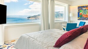 Une chambre d’hôtel équipée d’un lit, d’un bureau, d’une chaise, d’une télévision et d’une grande fenêtre donnant sur la mer et les falaises.