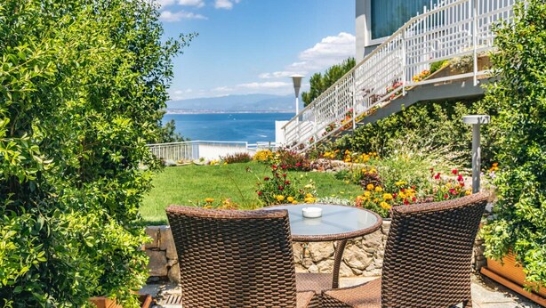 Une terrasse dotée de chaises en osier et d’une table en verre, donnant sur un jardin aux fleurs multicolores et offrant une vue sur la mer.