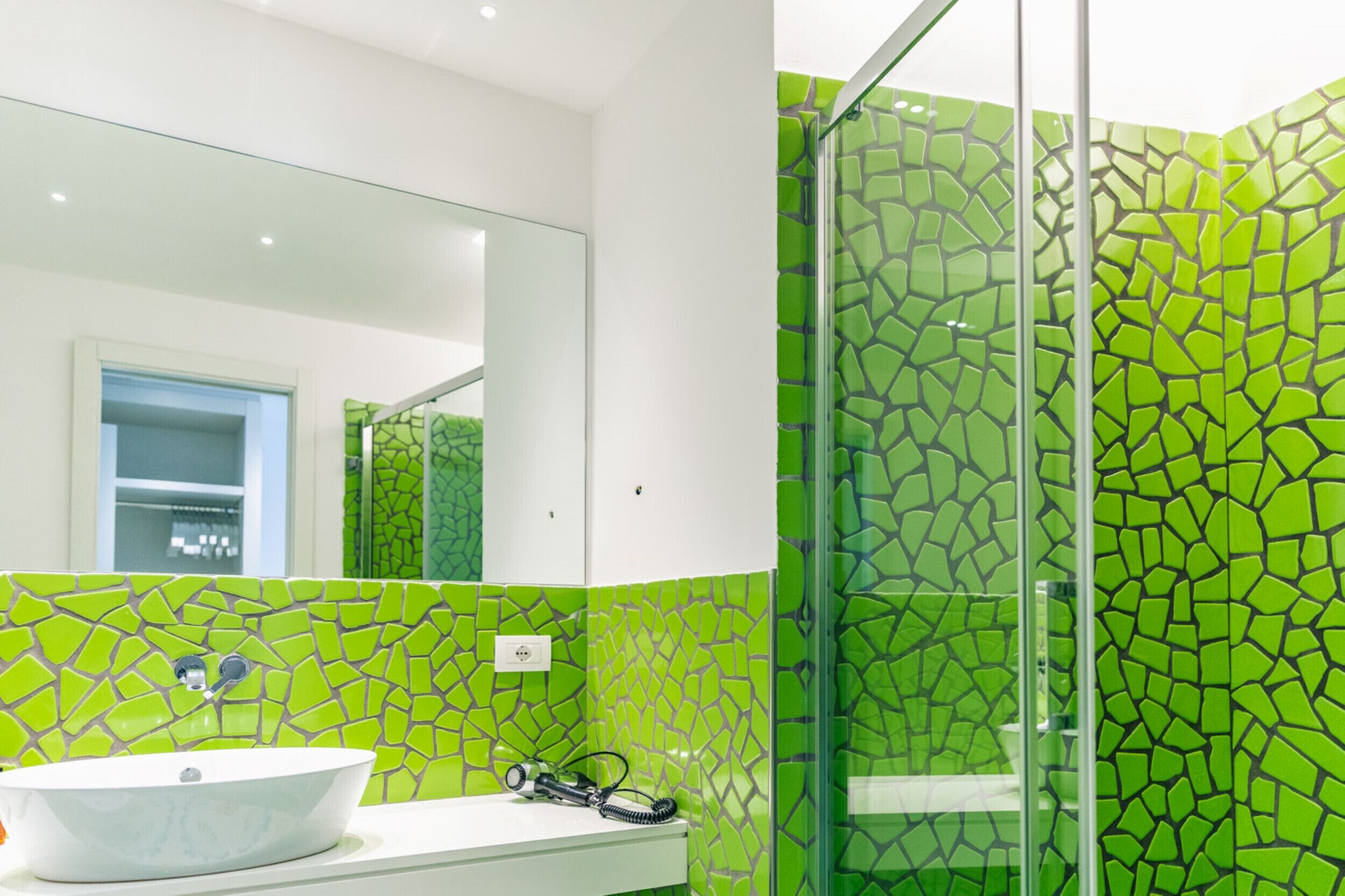 A bathroom with a green mosaic tile wall, a white sink, and a glass shower enclosure.
