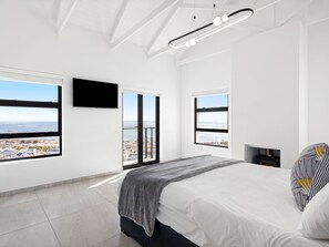 A modern bedroom with a large bed, a TV mounted on the wall, and a view of the city through large windows.