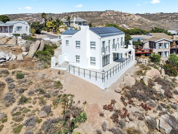 A modern two-story house with a flat roof and solar panels, surrounded by rocky terrain and sparse vegetation.